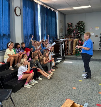 A woman directs a seated group of children holding books and papers, with a clock and blue curtains in the background.