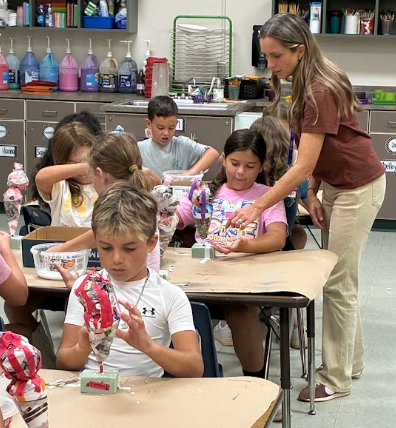 Classroom with students at tables, engaging in art projects. Teacher assists a child in front, surrounded by colorful supplies on shelves.
