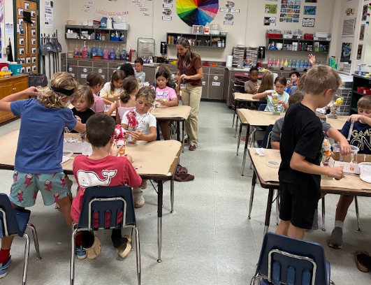 A classroom with children seated at tables, some standing. A woman in a brown shirt stands near a table.