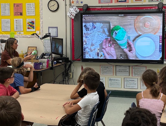 Classroom with a teacher showing a presentation on a monitor. Students sit at a table and on chairs.