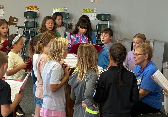 A classroom where children in different colored shirts gather around a piano with music sheets.