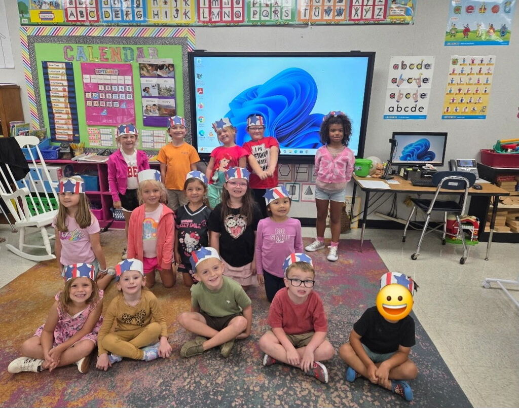 A classroom with students wearing hats; some are sitting on the floor and others are standing.