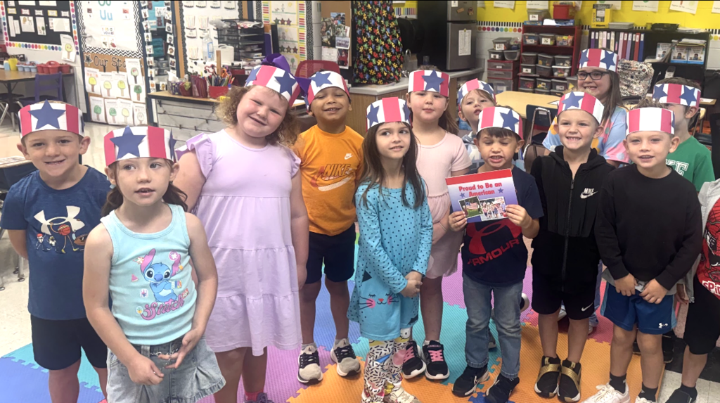 A classroom with children in patriotic hats, some holding books. Background includes shelves and a Christmas tree.