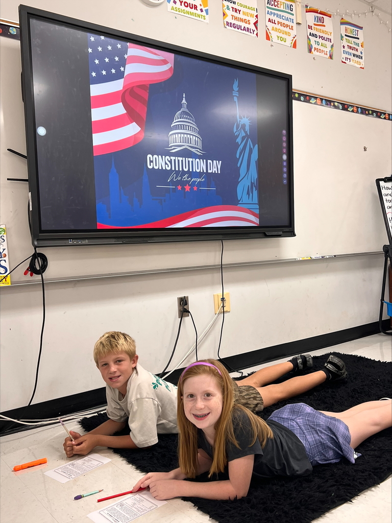 Two children lie on a rug, smiling, in front of a large monitor displaying a Constitution Day graphic.