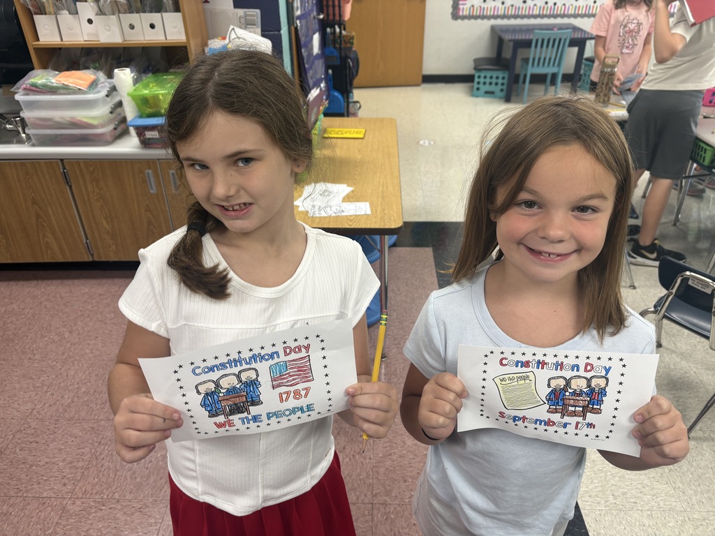 Two girls in a classroom hold up signs that say "Constitution Day." One wears a white shirt and red skirt, and the other a white shirt.