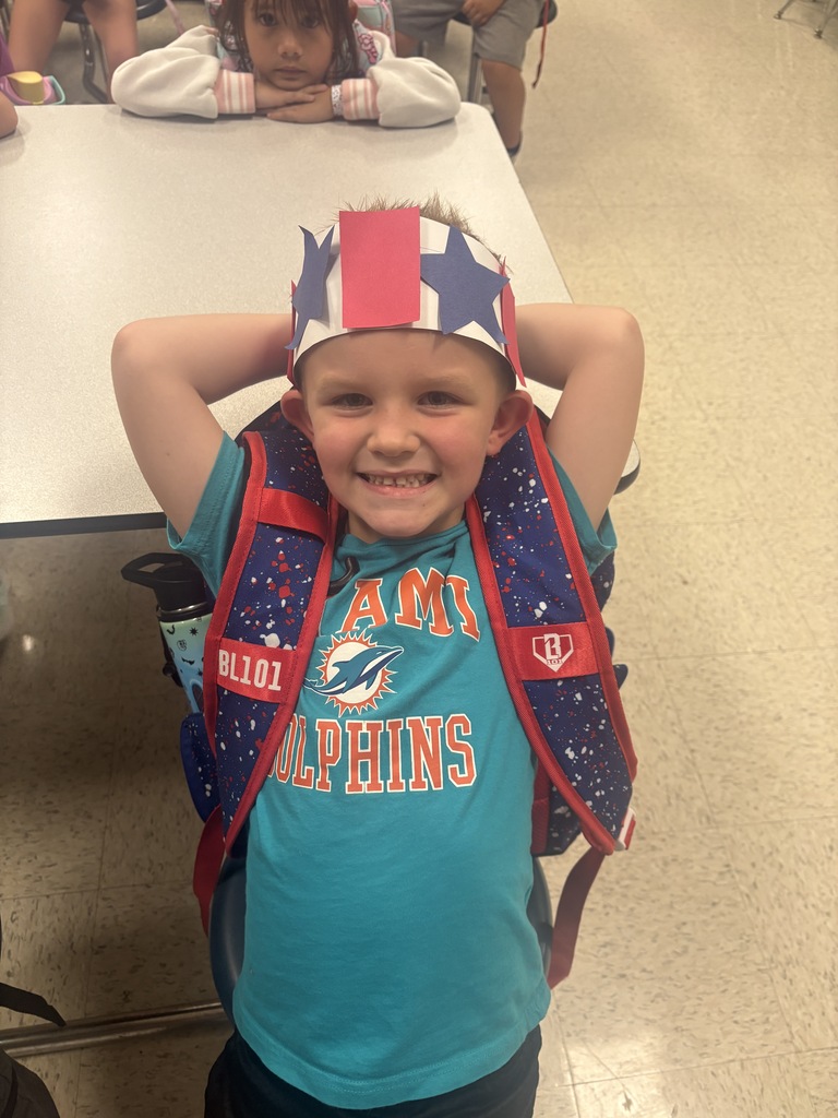 A boy wearing a Miami Dolphins backpack and a hat with stars smiles at the camera.