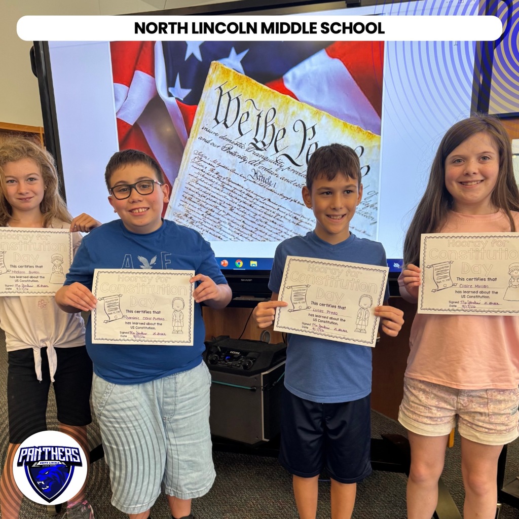 Four students stand holding certificates in a classroom with a large screen displaying "We the People."