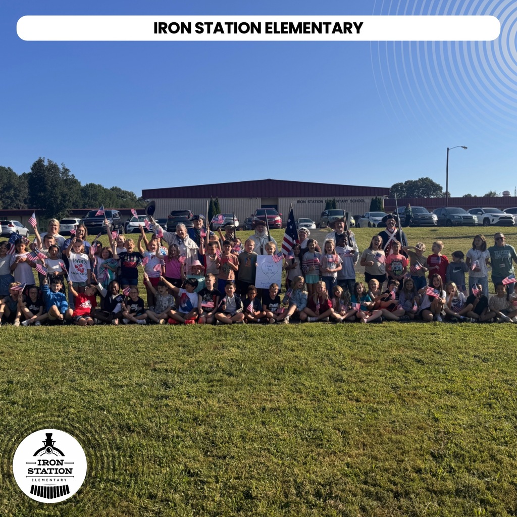 Many children and adults in a grassy field, holding flags. A sign at top reads "Iron Station Elementary".