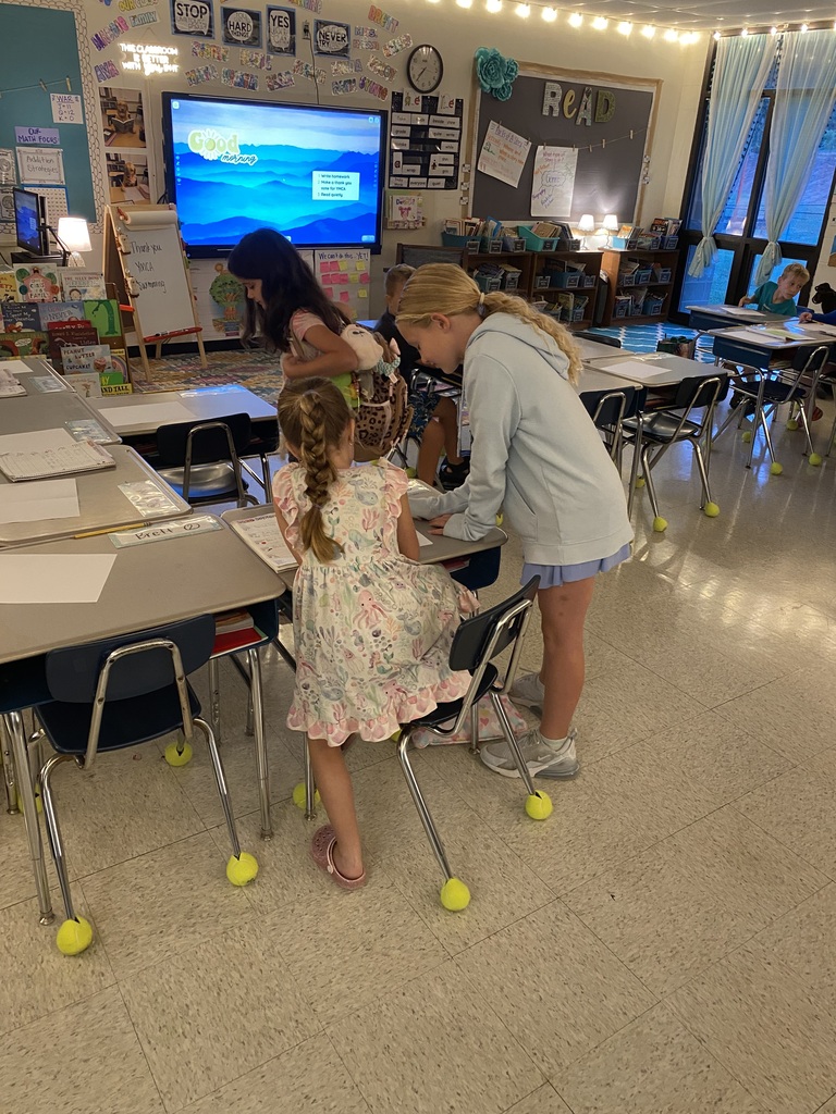 A teacher helps a student seated on a chair in a classroom with a monitor, tables, chairs, and a bookshelf.