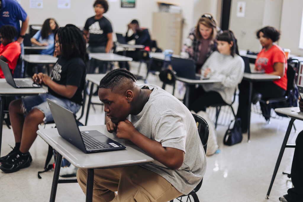 A classroom setting with students using laptops; a person in white sits on a chair at the front.