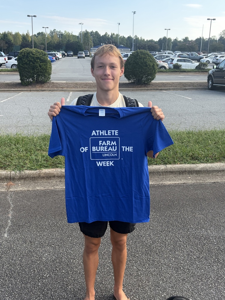 A young man holds up a blue t-shirt, reading "Athlete of the Farm Bureau Week," in a parking lot with trees and parked cars.