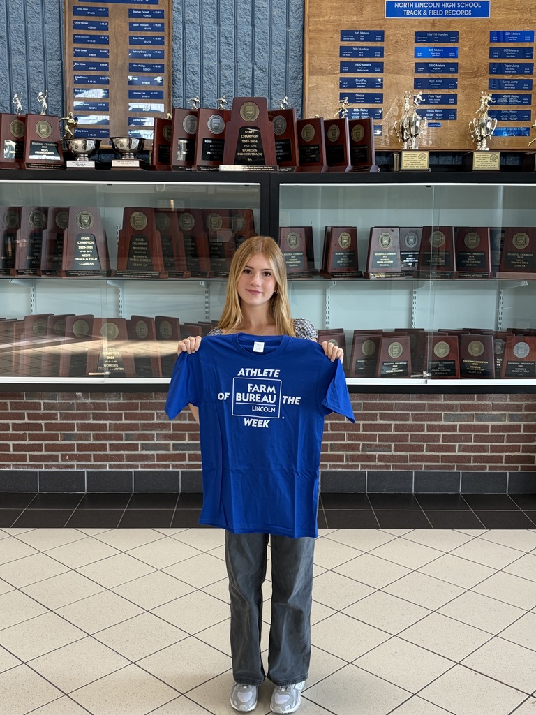 A girl stands in a hallway, holding a blue shirt, with a trophy case and brick wall behind her.