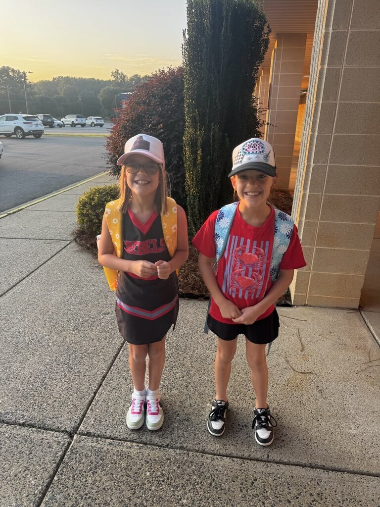 Two children wearing hats, backpacks, and sneakers stand on a sidewalk with a bush behind them.