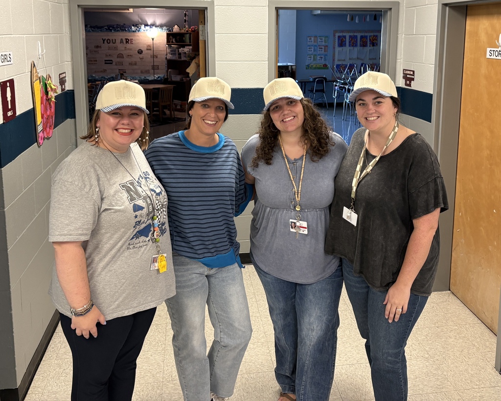 Four women, all wearing hats and name tags, stand together in a hallway, two doors visible behind them.