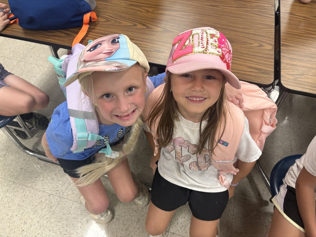 Two girls in school uniforms, one with a blue shirt and cap, the other with a pink hat, standing next to each other with backpacks.