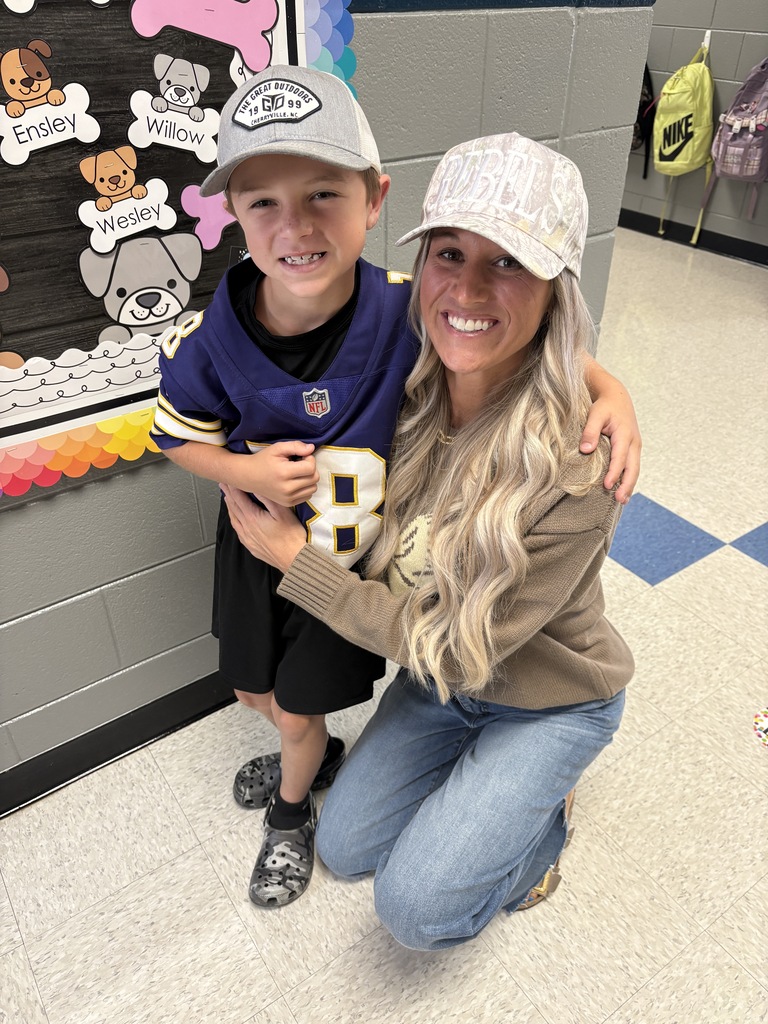 A woman in a beige sweater and jeans poses with a child in a blue jersey.