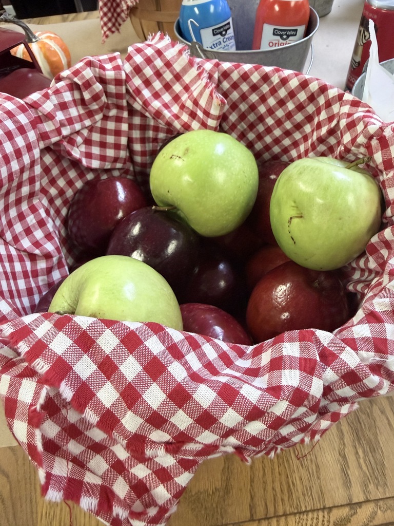 A basket filled with green and red apples, covered in a red and white checkered cloth, on a wooden table.