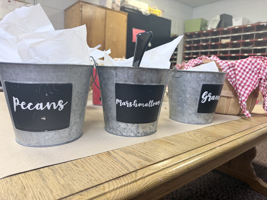Three buckets filled with paper bags labeled "Pecans", "Marshmallows", and "Grains" sit on a table. A wooden cabinet and shelves are in the background.
