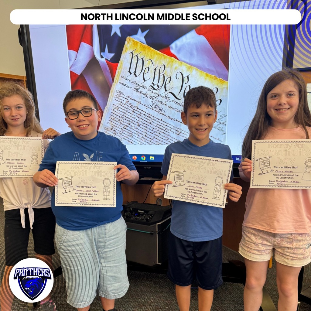 Four children stand together, each holding a certificate, in a classroom. A large screen behind them displays the American flag.
