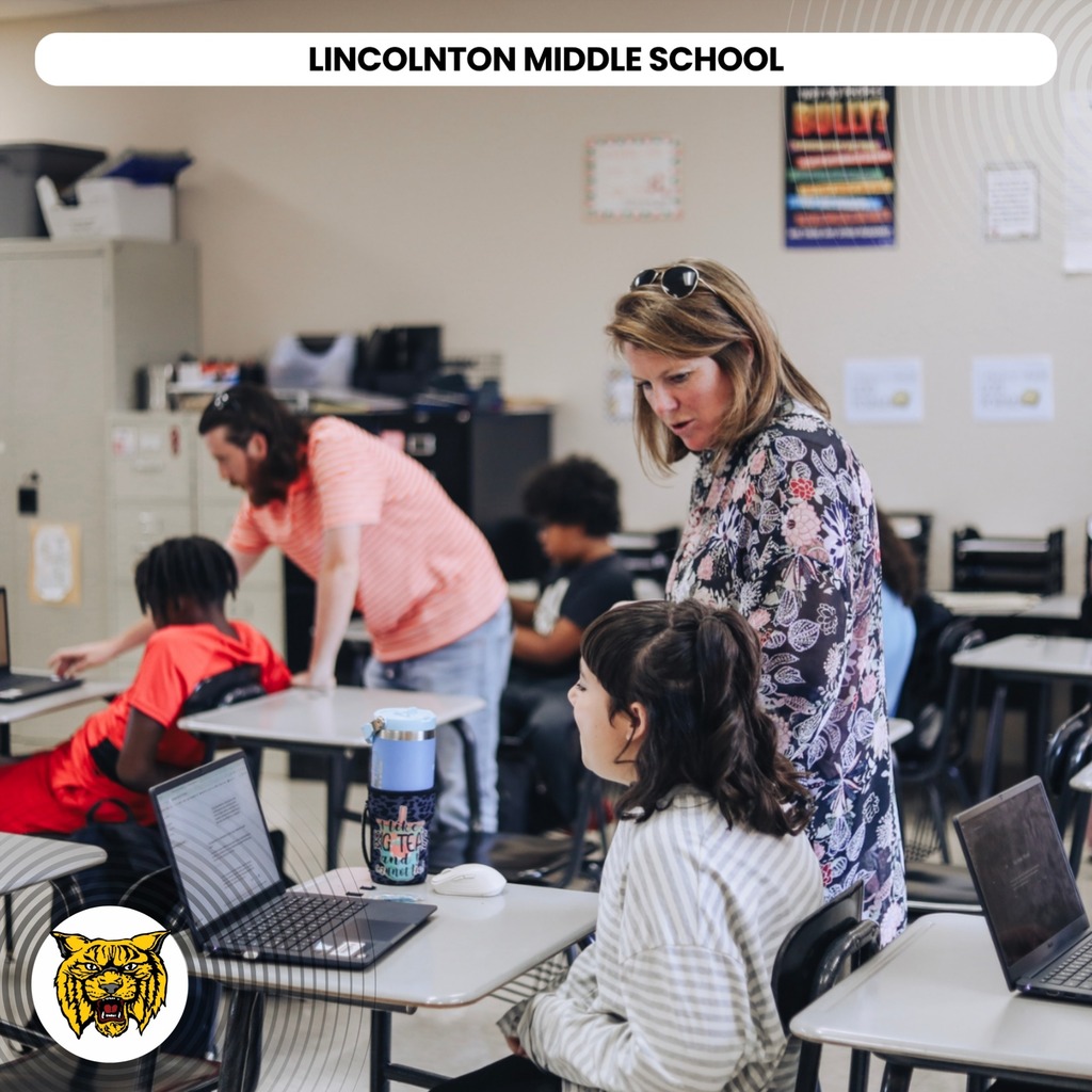 A classroom with students using laptops. A teacher assists a girl, and another teacher helps a boy.