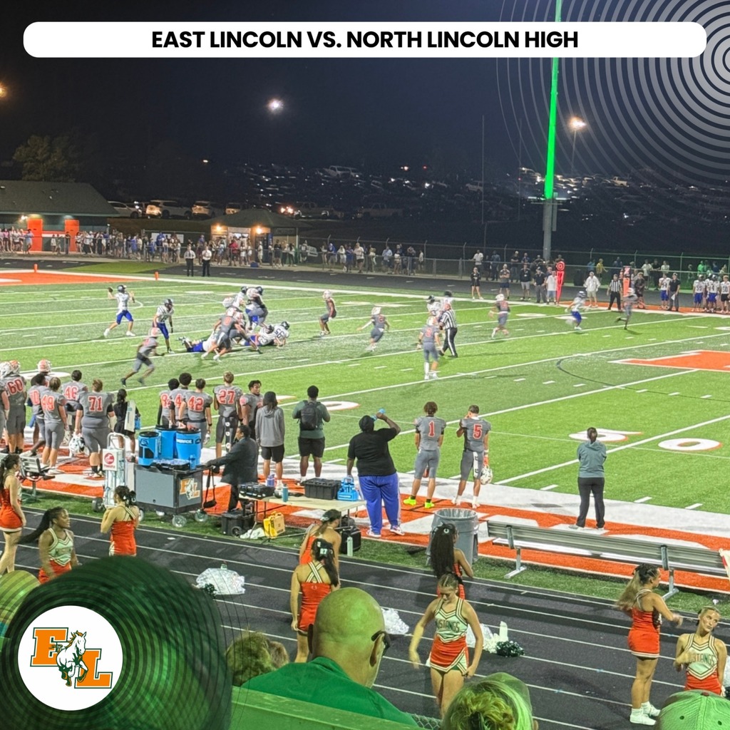 Football game at night, players on field, spectators in stands, cheerleaders on sidelines, large banner overhead.