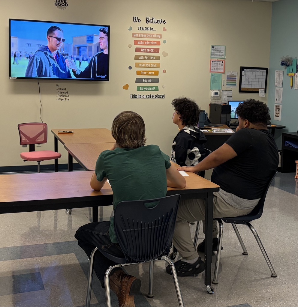 Three individuals seated in a classroom with a monitor showing a person on screen. Bright walls with posters and a calendar.
