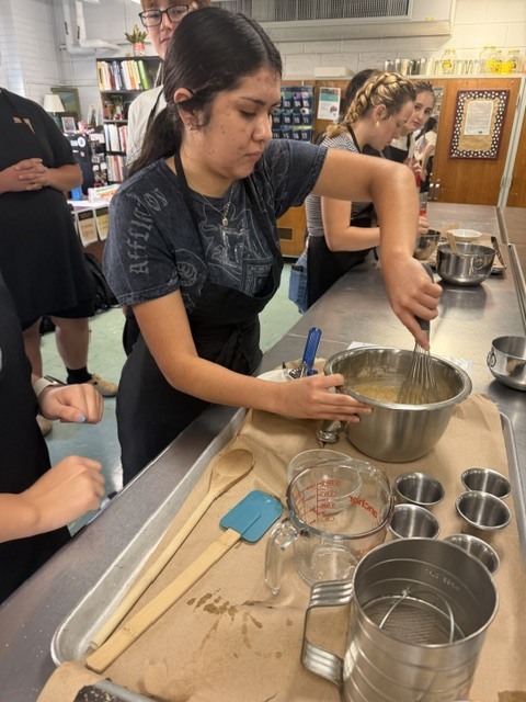 People in a kitchen. A woman mixes in a bowl, with others working nearby. A table holds utensils.