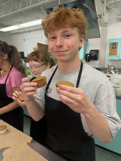 A young man in a gray t-shirt and black apron holds cookies in a classroom kitchen.