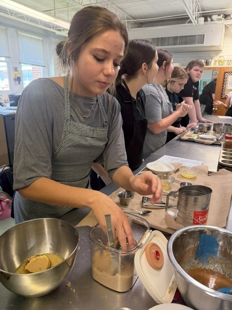 Woman in apron preparing food in a kitchen with other people working on the countertop.