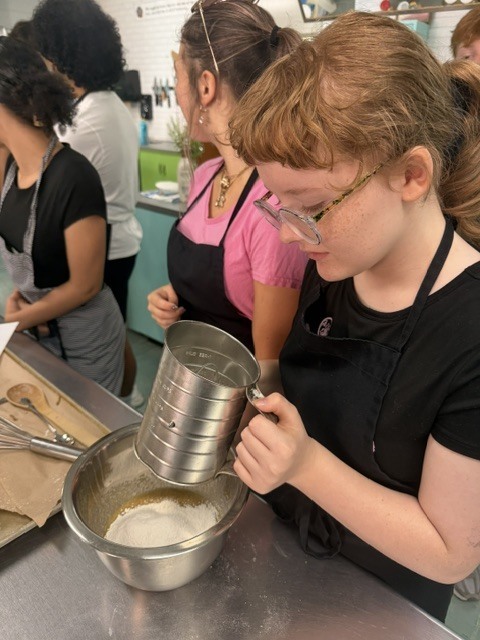 A woman in a black shirt and apron pours ingredients into a bowl in a kitchen.