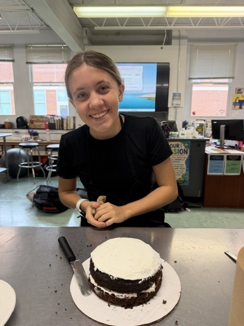 A woman smiles while standing behind a table, frosting a cake. A large room with tables and chairs is in the background.