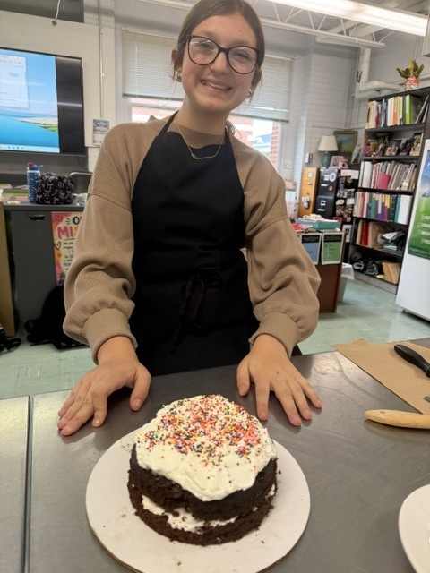A smiling person in a brown sweater and apron stands behind a table with a chocolate cake.