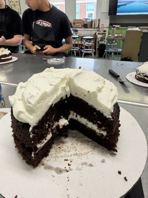 A three-layer chocolate cake with white frosting sits on a white plate. Behind it, two people work at a table.