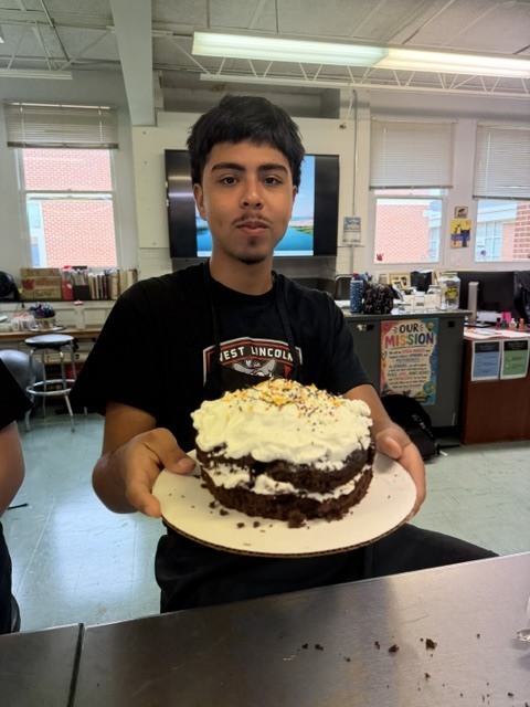 A person wearing a black shirt is holding a white plate with a chocolate cake and white cream.