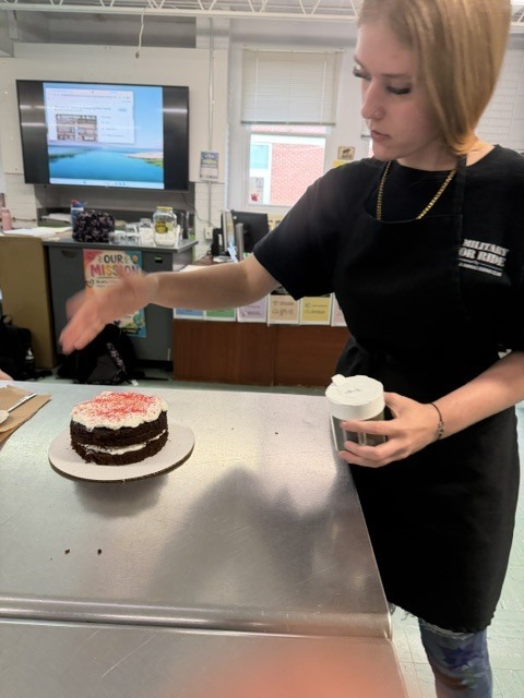 Woman in black shirt decorates cake with pink frosting. A monitor and desk are in the background.