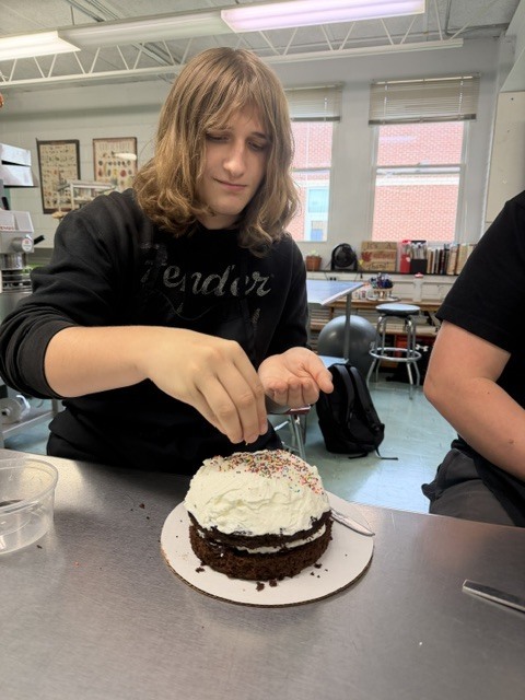 A person decorates a cake with frosting and sprinkles in a room with tables and chairs.