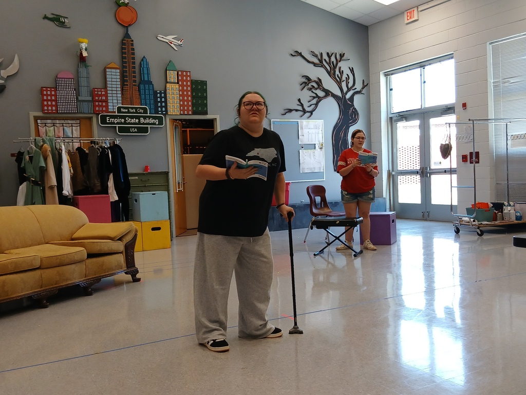 A student holding a script and leaning on a walking cane stands in the foreground during a drama rehearsal while another student reads a script in the background.