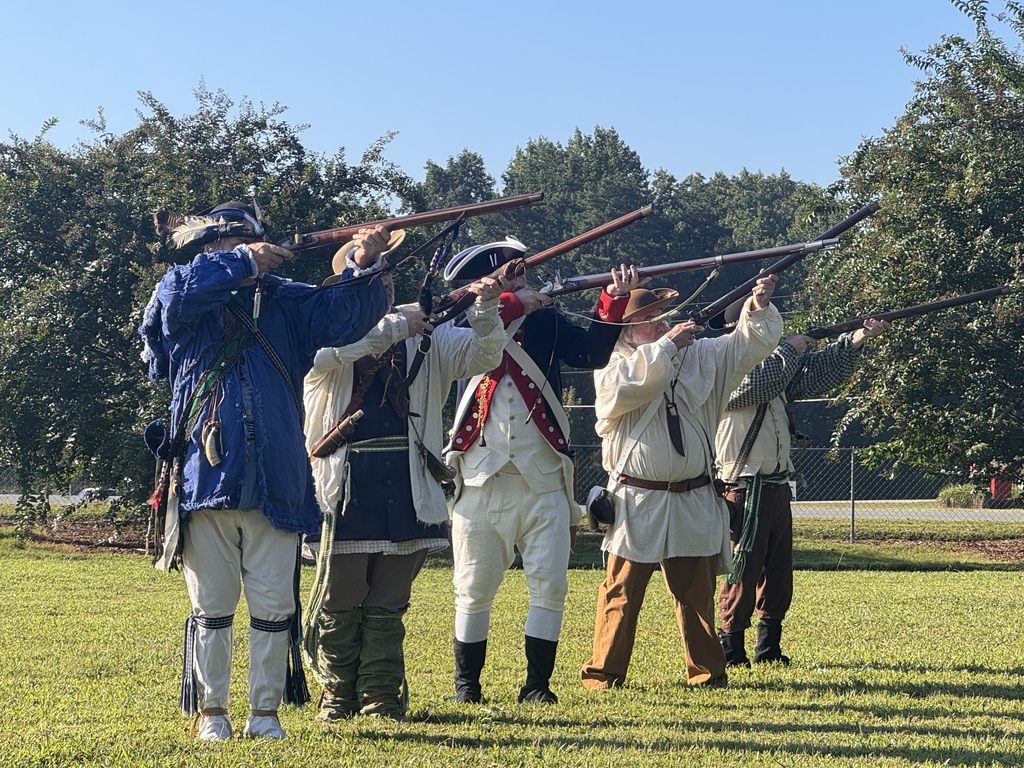 Five historical re-enactors in Revolutionary War-era clothing stand in a line on a grassy field outdoors, each aiming a musket upward into the sky.