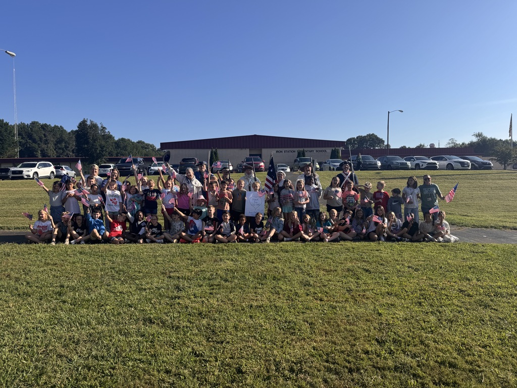 A large group of elementary students and adults, including re-enactors in historical military attire holding muskets and flags, posing together outdoors on a lawn in front of Iron Station Elementary School. Many students are sitting or standing on a paved walkway, waving small American flags.