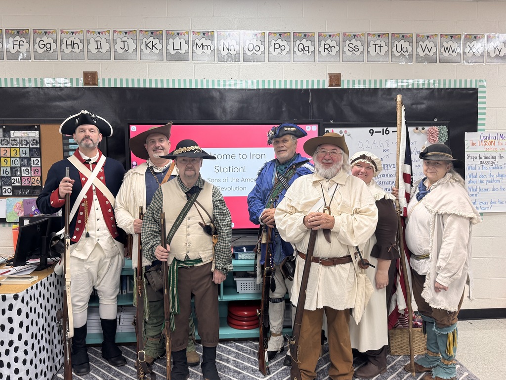 Seven historical re-enactors dressed in Revolutionary War-era attire stand in a classroom in front of a digital display reading "Welcome to Iron Station! Sons of the American Revolution." The men and women are wearing period-appropriate tricorn hats, coats, waistcoats, and dresses, with several holding muskets and an American flag.