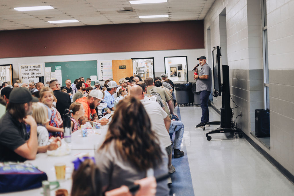 A man speaking into a microphone at the front of a crowded school cafeteria filled with adults and young children seated at long tables. A poster on the back wall reads, "Build a Team so Strong That NO one Can point..."