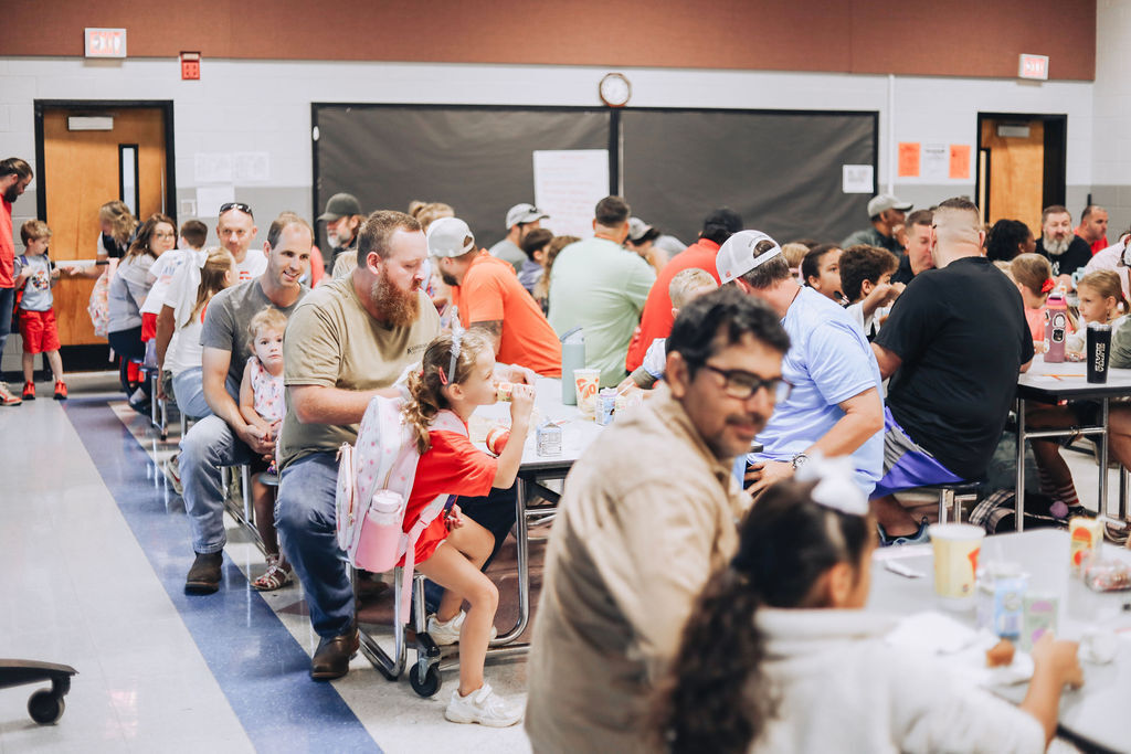Elementary students and family members sitting along long cafeteria tables during a school event, eating breakfast and talking in a full cafeteria.
