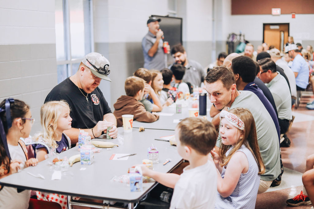 Adults and elementary school students sitting together at long cafeteria tables during a school event. In the foreground, children sit across from adult figures, eating breakfast items and chatting, while a speaker with a microphone addresses the room in the background.