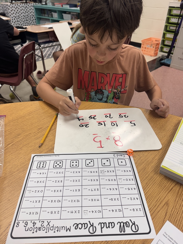 An elementary student wearing a Marvel shirt sitting at a wooden classroom table and writing numbers on a small dry-erase board with a marker. Below the whiteboard, a printable multiplication activity card titled "Roll and Race Multiplication" lies on the desk alongside a yellow pencil.
