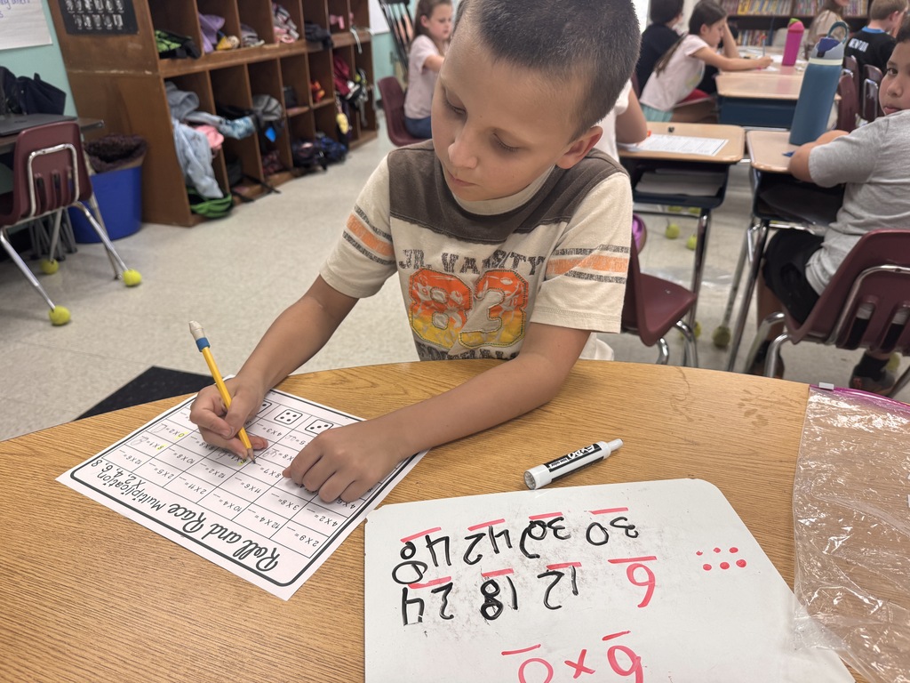 An elementary student sitting at a classroom desk completing a math activity titled "Roll and Race Multiplication." The student holds a pencil over a worksheet while a small whiteboard on the desk displays written numbers and math equations. Other students are working at desks in the background.