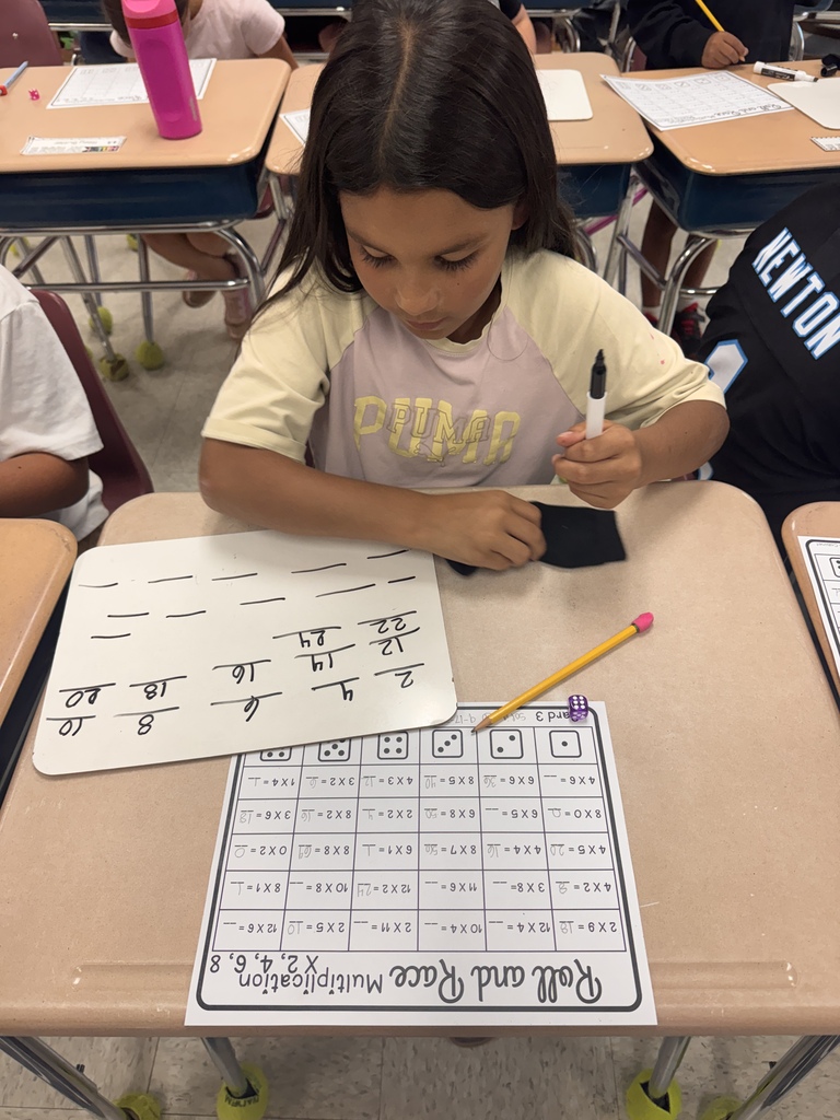 An elementary student sitting at a classroom desk, holding a marker over a small whiteboard filled with written numbers. In front of the whiteboard is a multiplication game sheet titled "Roll and Race Multiplication" along with a yellow pencil and a purple die.