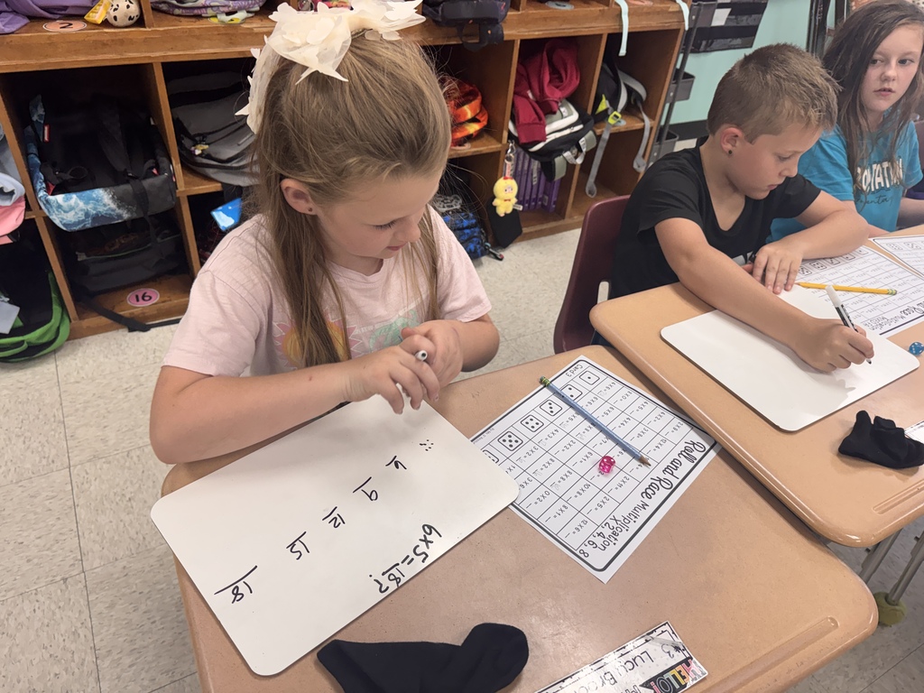 An elementary student with a white hair bow sitting at a desk and working on a math problem using a small whiteboard and marker. On the desk next to the whiteboard, a game sheet titled "Roll and Race Multiplication" and dice are visible. Another student works at an adjacent desk in the background.