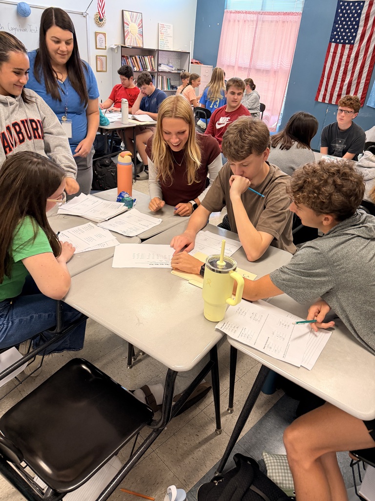Several students are seated around tables in a classroom, engaged in a discussion, with papers and a yellow mug on the table.