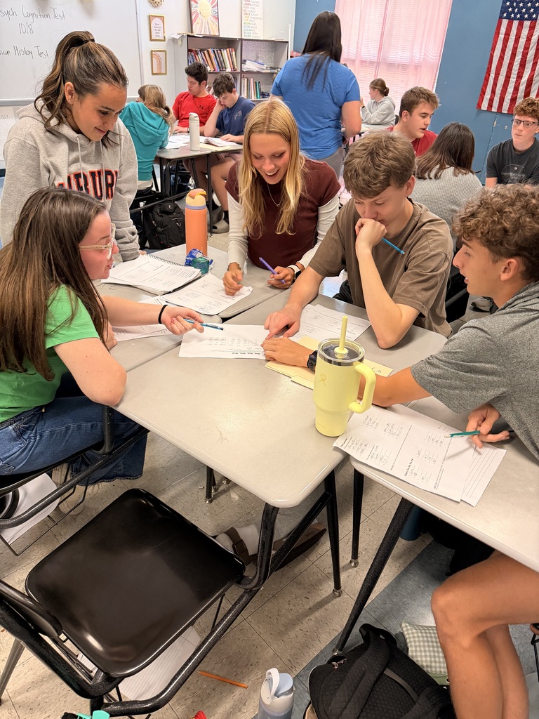 A classroom with students sitting at desks. Four students are engaged in conversation at a table with papers, a yellow mug, and a bottle.