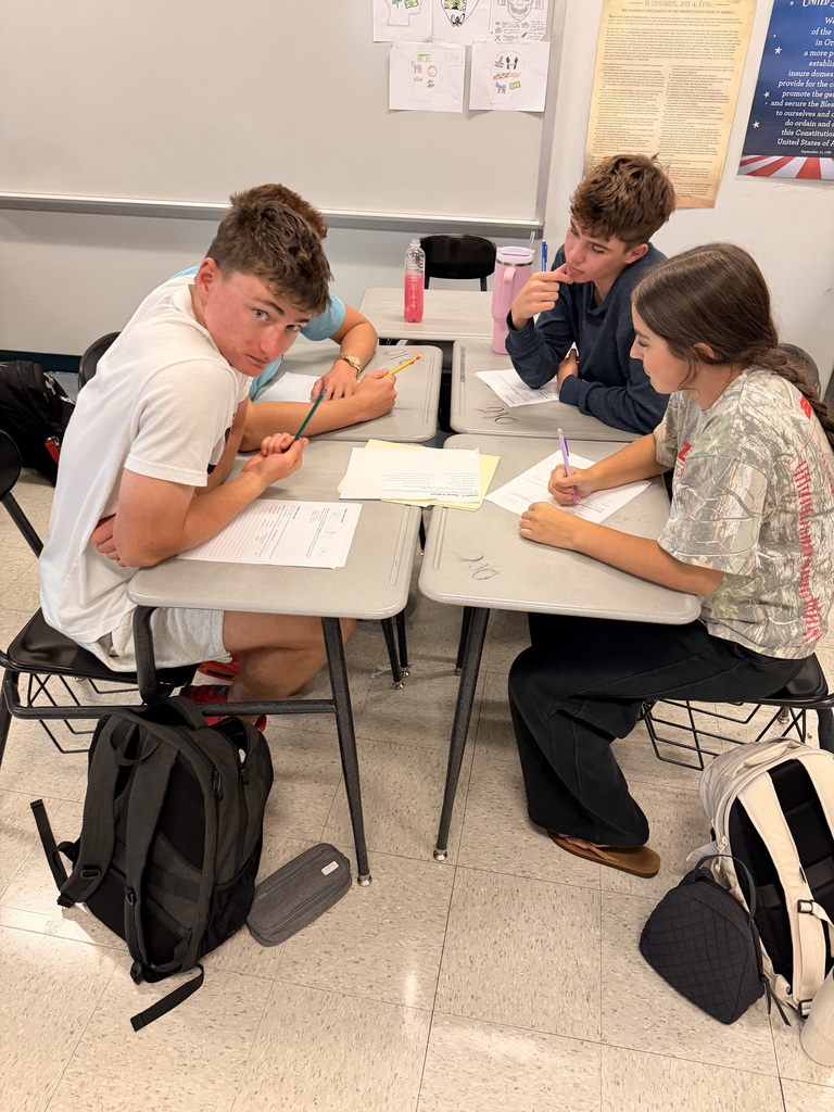 Four students sit at desks, each with papers and pens, in a classroom. Backpacks are placed near them.